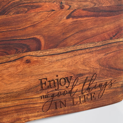 A close-up of a rustic wooden board with engraved text reading 'Enjoy the good things IN LIFE' on a white surface, with a white wall and power outlet visible in the background.