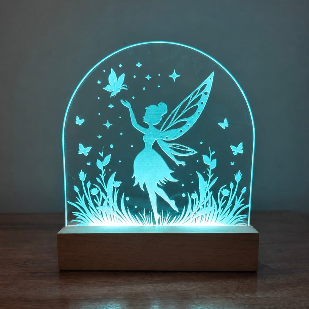 A fairy-themed 3D LED night light with a glowing blue silhouette of a fairy, butterflies, and grass on an arched acrylic panel mounted on a wooden base, set on a dark wooden table against a dark gray background.