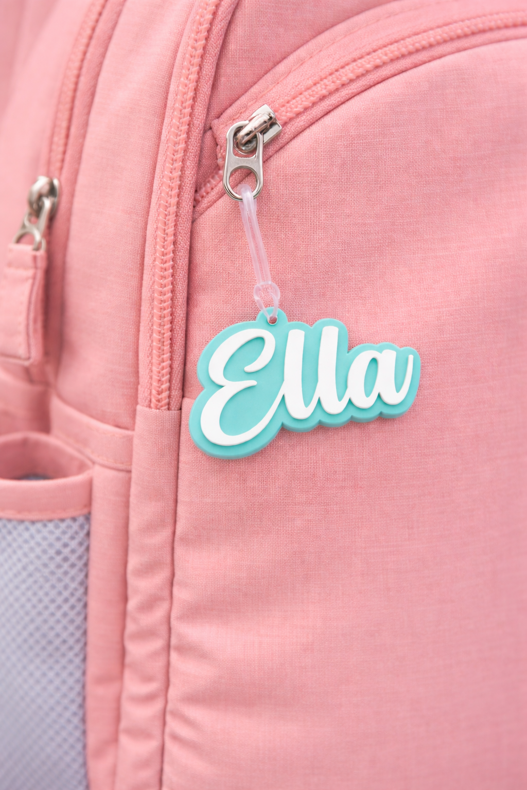 Aqua acrylic name bag tag reading Ella in script font clipped to a pink school bag zip