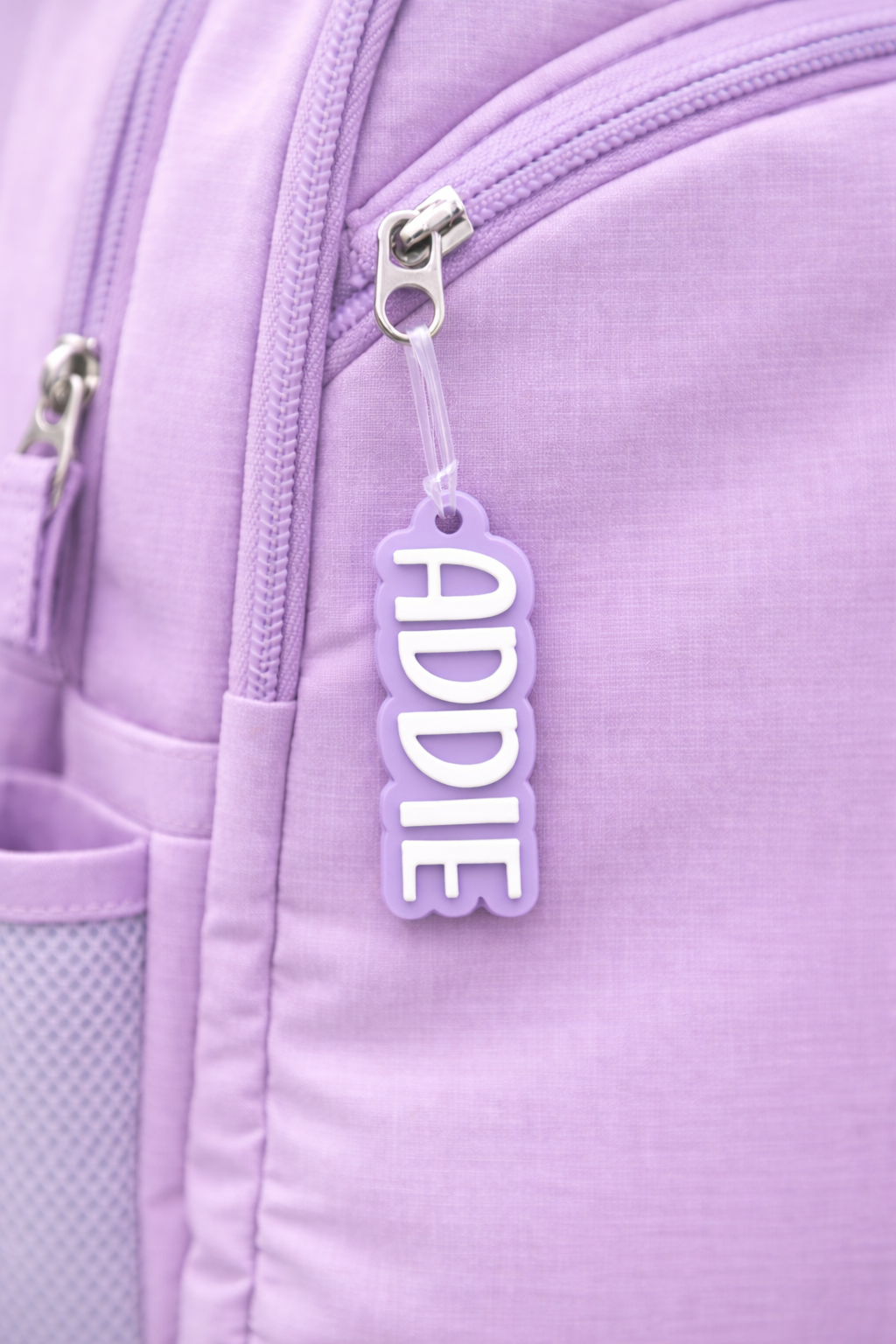 Purple acrylic name bag tag reading ADDIE in bold font on a lilac backpack zip